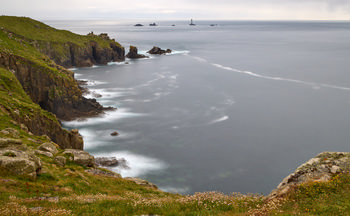 Long exposure longships This is a landscape photograph taken on a spring evening, capturing the rugged cliffs and sweeping grassy coastline of Cornwall. The image prominently features the sea with a smooth, misty appearance achieved through long exposure, accentuating the motion of the waves against the rocks. In the distance, the Longships lighthouse rises above the surrounding sea and rocky outcrops, serving as a notable landmark. The cliffs, covered in green vegetation, dominate the foreground, framing the expansive view towards the Longships lighthouse and the surrounding waters.